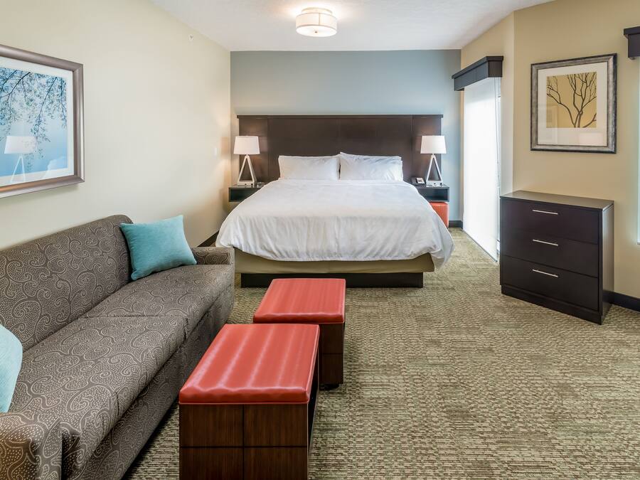 Quarto de hotel com cama, sofá, dois pufes vermelhos, mesa de cabeceira com abajur, penteadeira e duas imagens emolduradas nas paredes.
