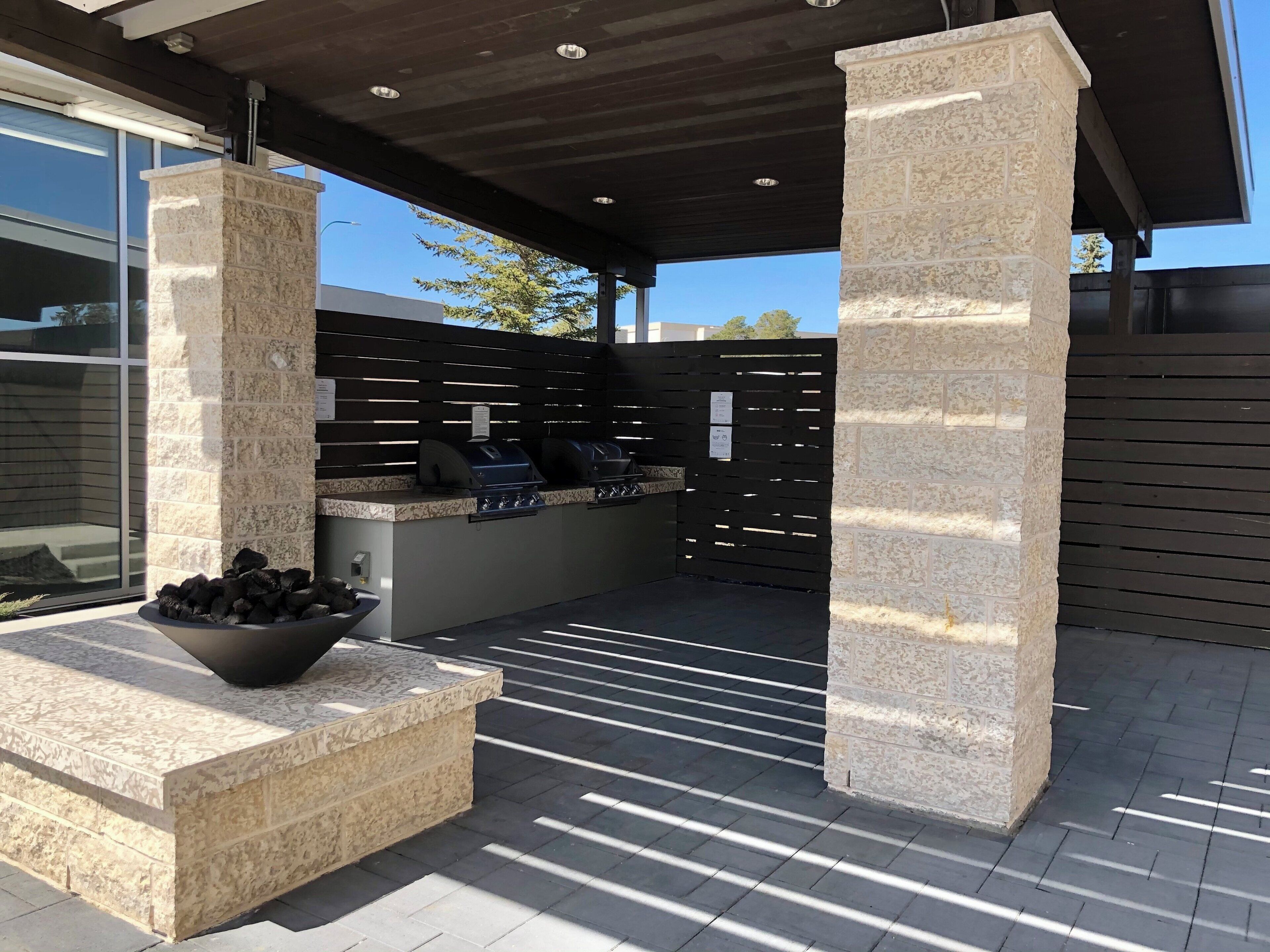 An outdoor patio area with a stone bench, a grill station, and a view of a modern building.