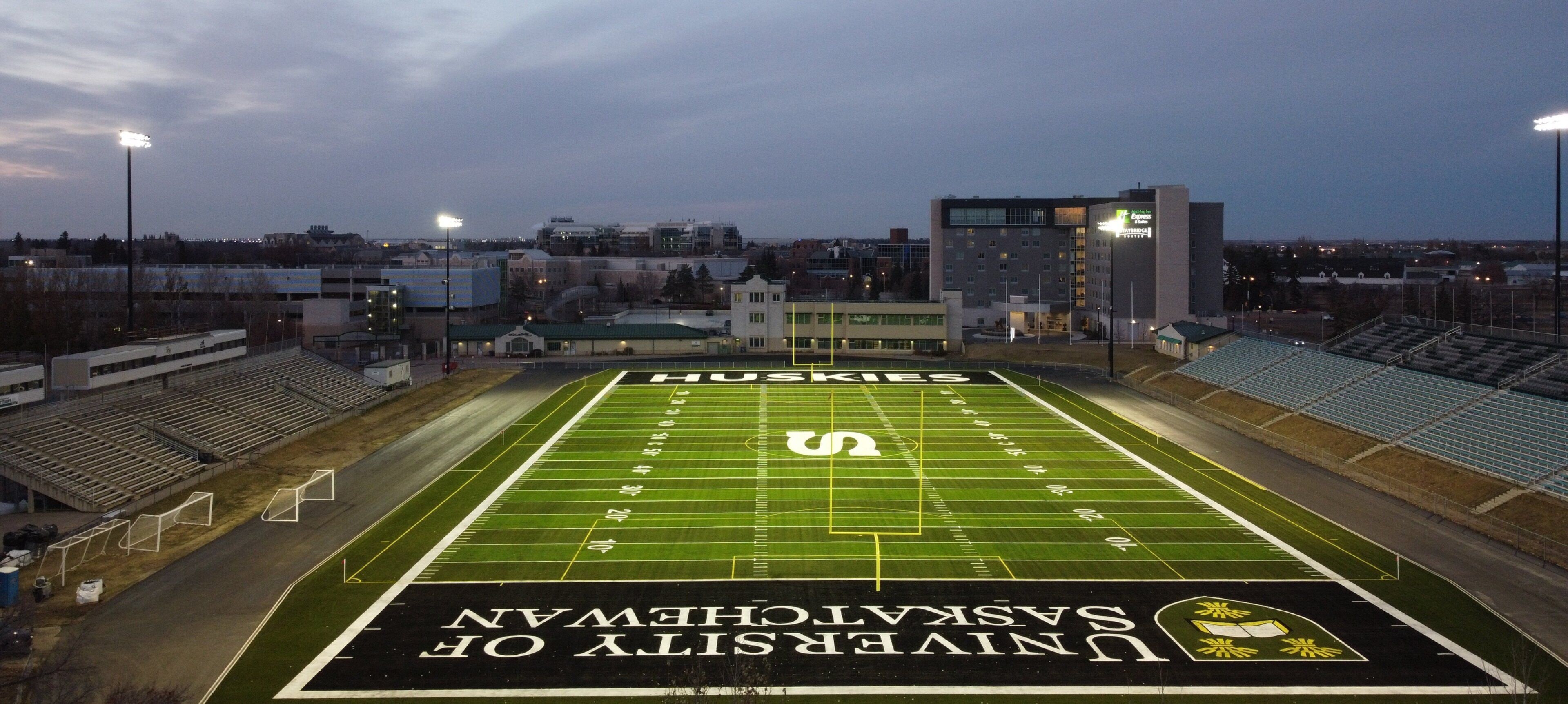 A football field with 'Huskies' and 'University of Saskatchewan' markings, surrounded by bleachers and buildings.