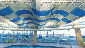 An indoor swimming pool with a wavy ceiling design, clear glass walls, and a seating area with chairs and tables.