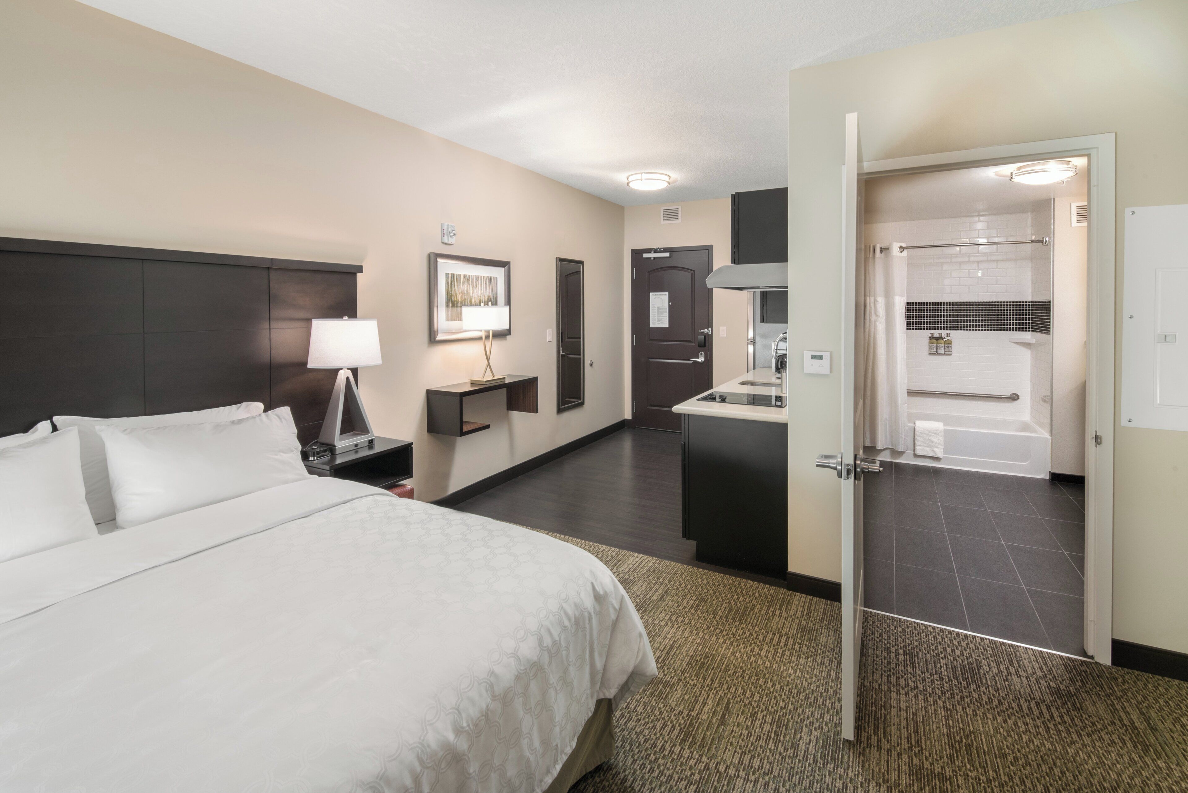 A hotel room with a bed, bedside table, lamp, artwork, and a view of the bathroom.