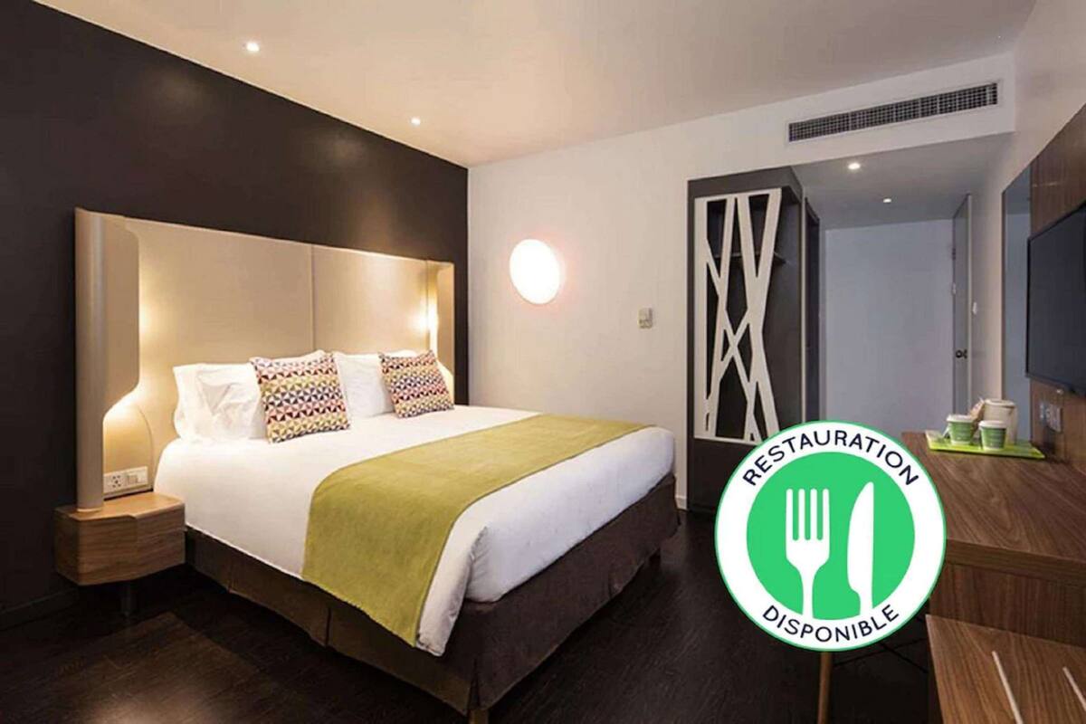 Ein modernes Hotelzimmer mit einem großen Bett, Nachttischen, einer grünen Tagesdecke und einem Schild, das die Verfügbarkeit eines Restaurants auf dem Gelände anzeigt.