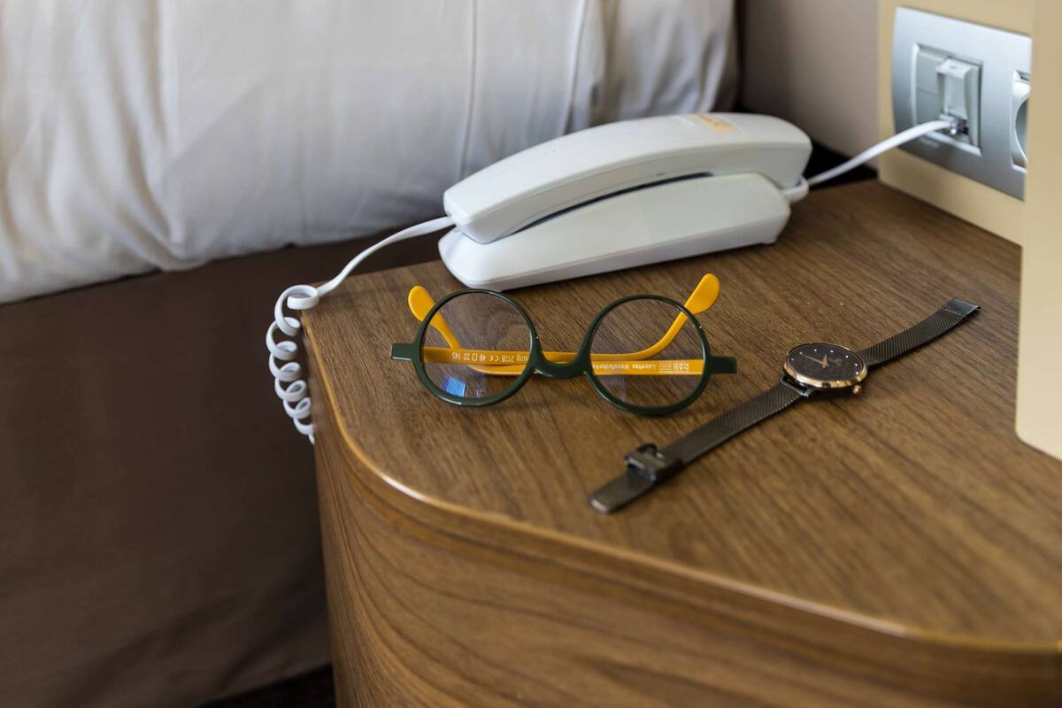 Ein ordentlich bezogenes Bett mit weißer Decke und Kissen, ein Nachttisch mit Telefon, Brille und Armbanduhr sowie eine Wandlampe.