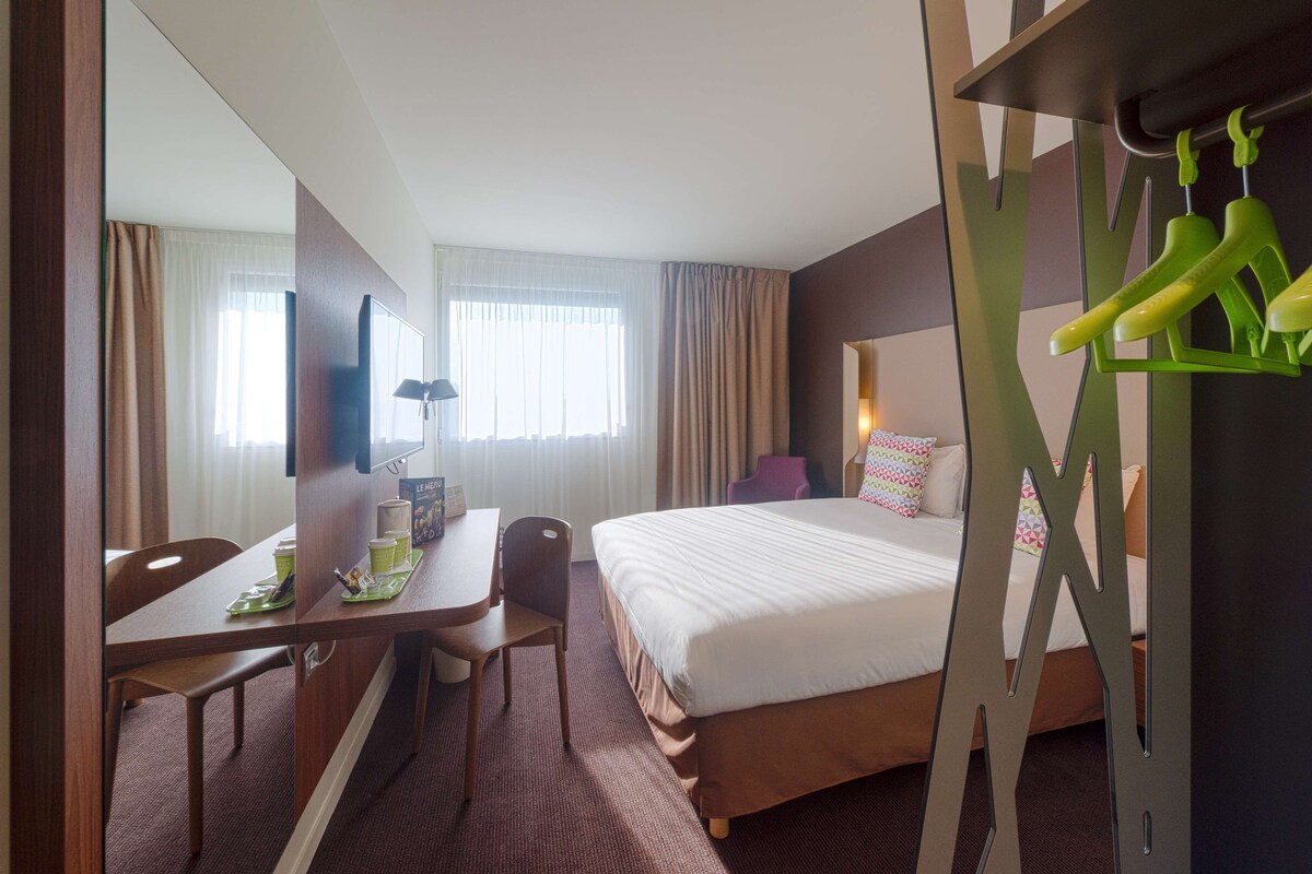 Ein Hotelzimmer mit Bett, Schreibtisch, Stuhl, Fernseher und einem Kleiderschrank mit grünen Kleiderbügeln.