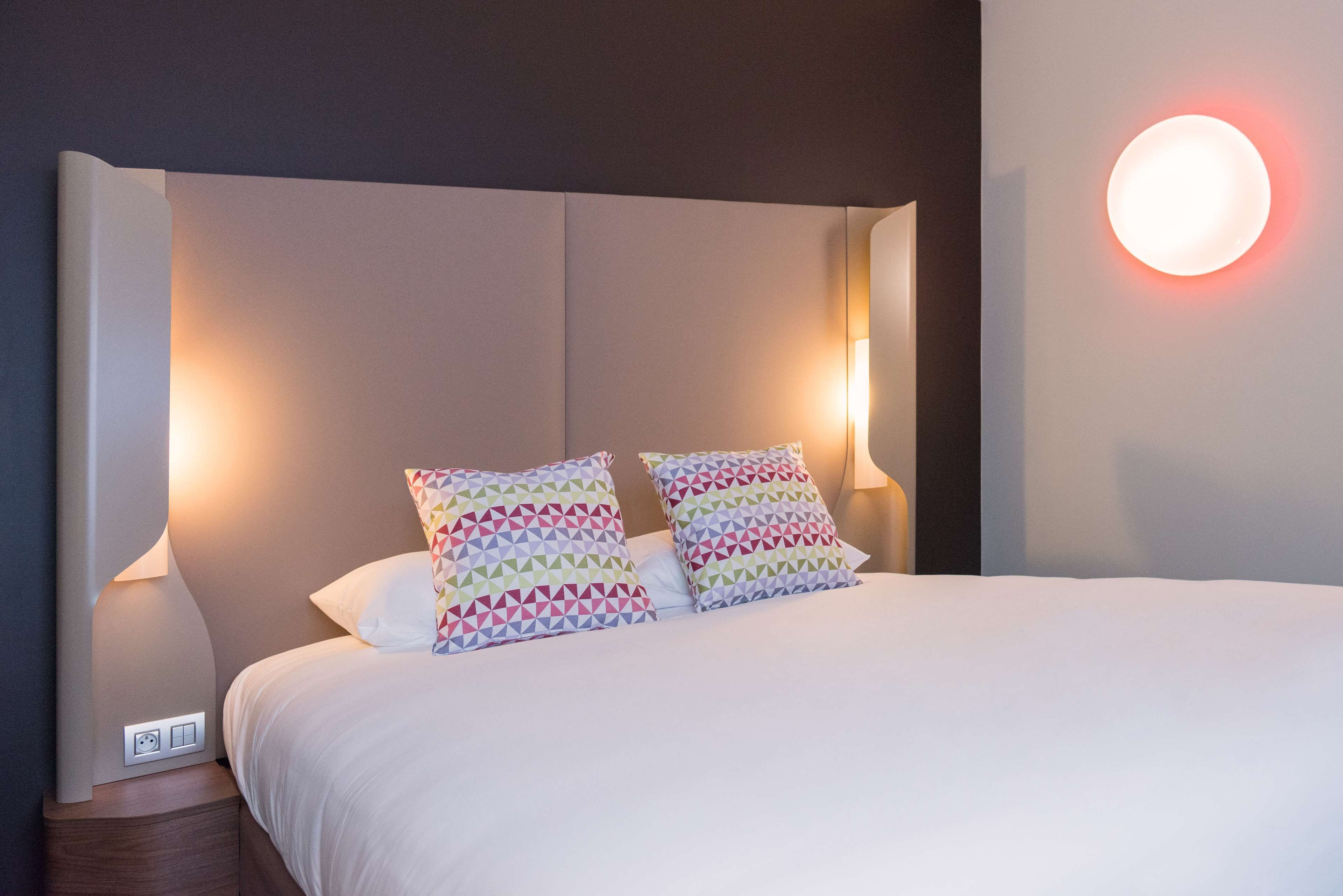 A neatly made bed with patterned pillows, a bedside table with a lamp, and a wall-mounted light.