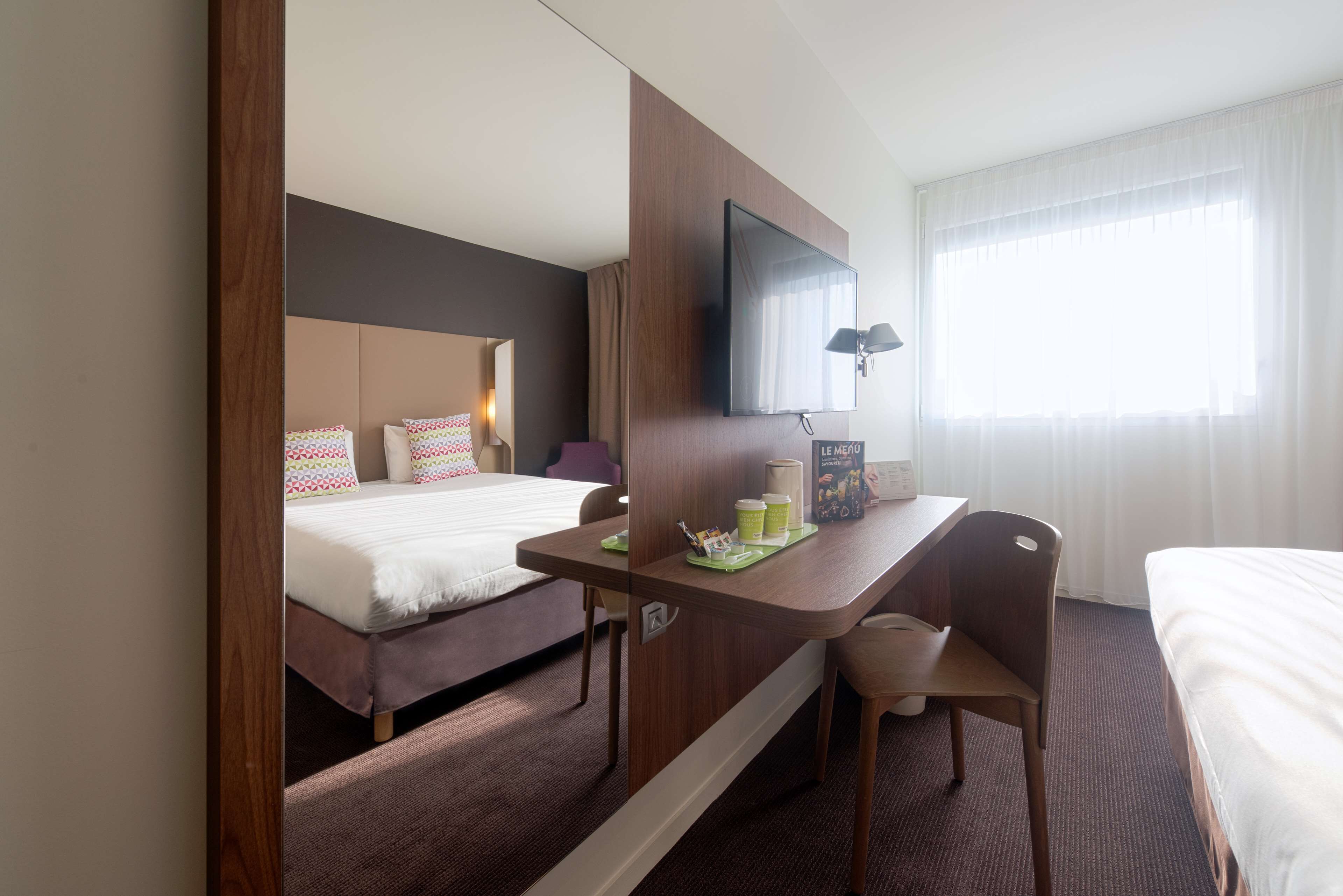 A hotel room with a bed, a desk with a chair, a flat-screen TV, and a window with curtains.