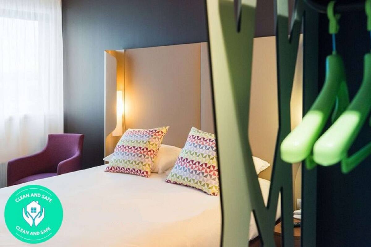 Ein Hotelzimmer mit einem Bett, bunten Kissen, einem Nachttisch, einer Lampe und einem grünen Sessel.