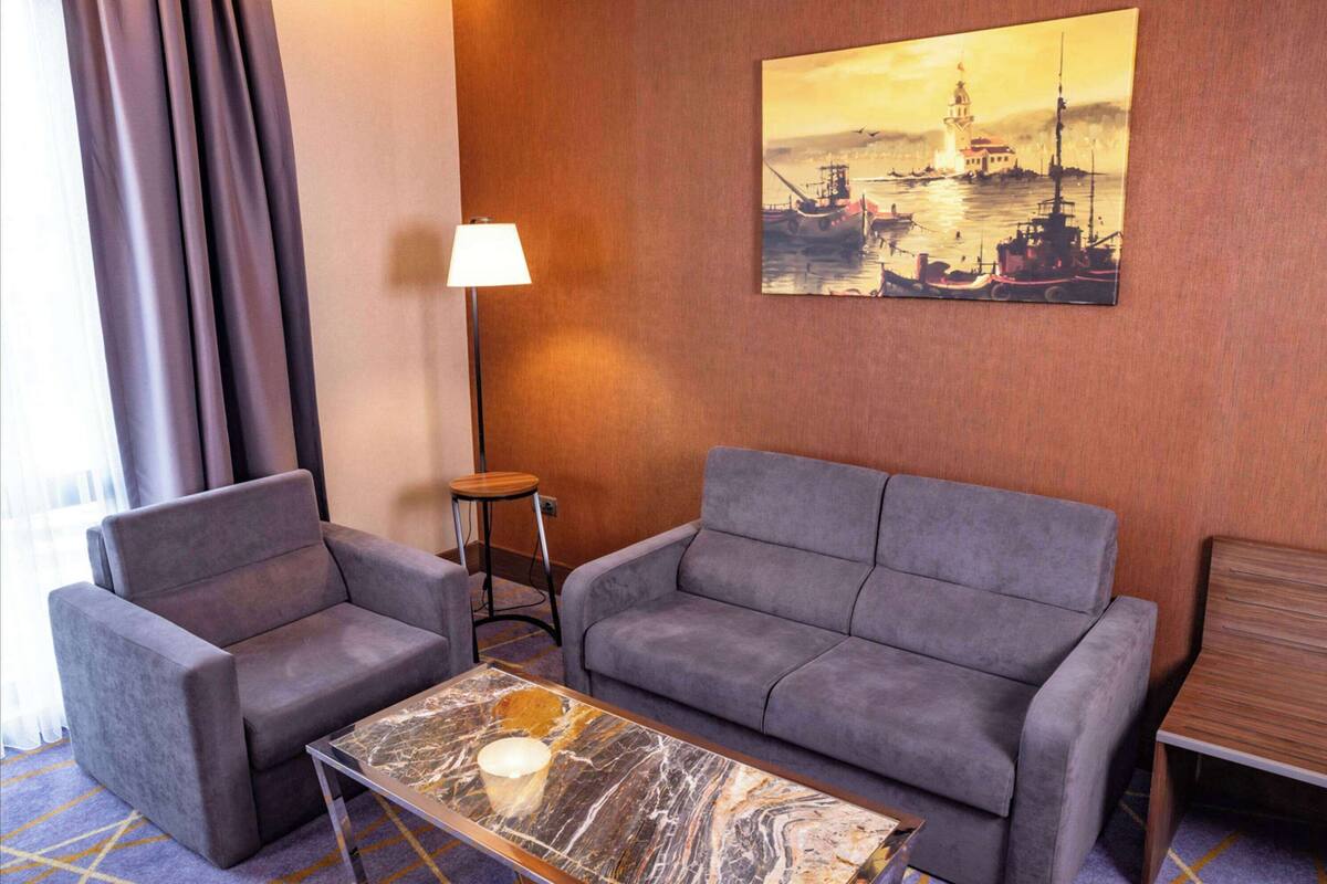 A hotel room with a grey sofa, a matching armchair, a marble coffee table, a lamp, and a painting of a coastal scene.