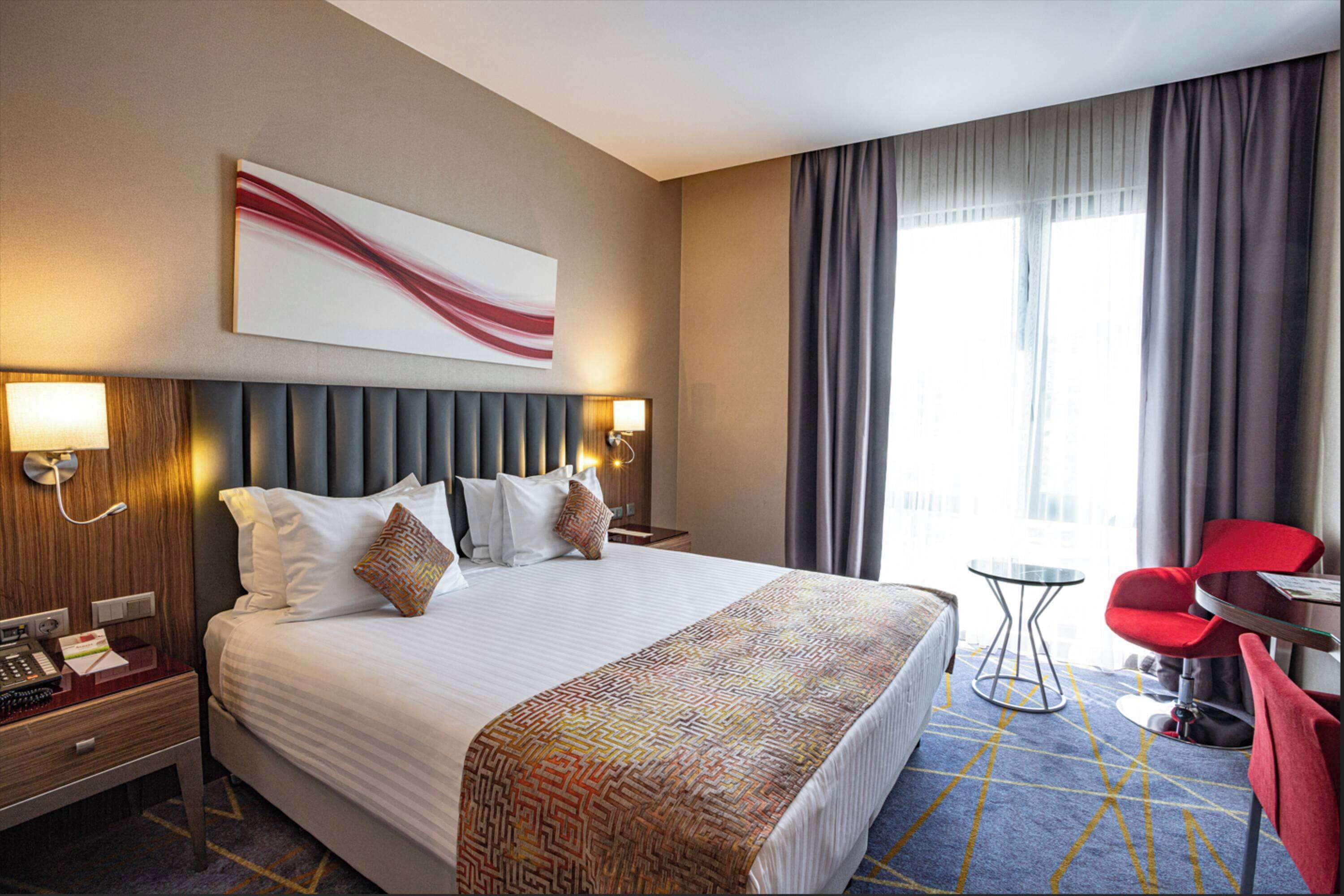 Une chambre d’hôtel avec un grand lit, une tête de lit dotée d’un luminaire, une table de chevet avec un téléphone et une lampe, une petite table ronde avec une chaise, et une fenêtre avec des rideaux.