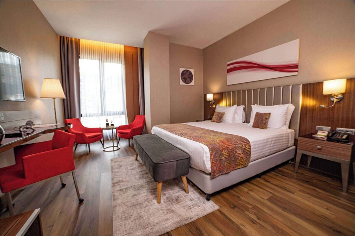 A hotel room with a large bed, a desk with a chair, a small table, and a red chair.