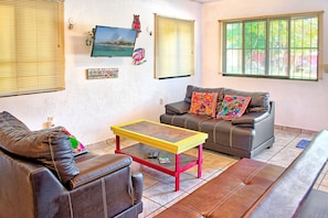 A living room with a brown leather sofa, a yellow and red coffee table, and a gray sofa with colorful pillows. - Caribe Suites (Puerto Morelos)
