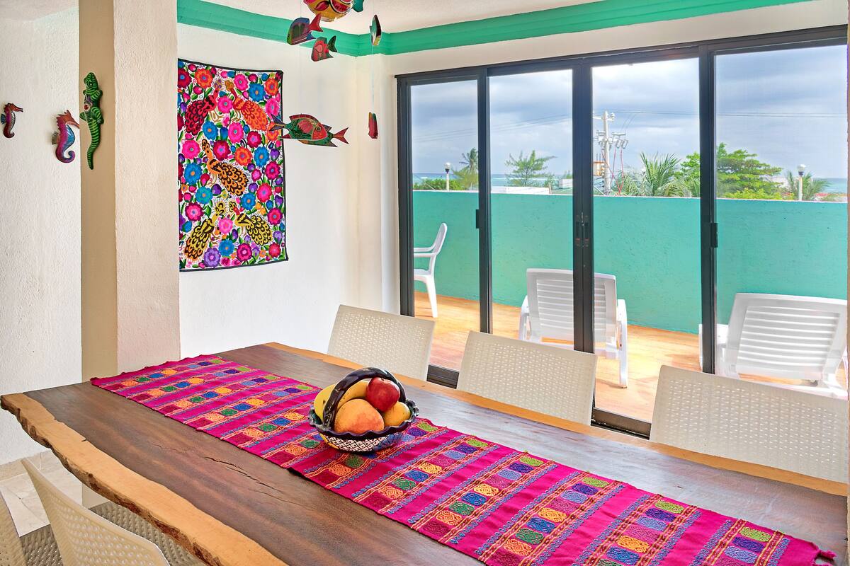 A dining area with a wooden table, colorful table runner, and a fruit basket.