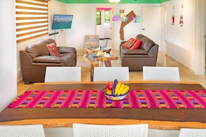 A living room with two brown leather sofas, a coffee table, and a dining table with a colorful placemat and a fruit bowl. - Caribe Suites (Puerto Morelos)