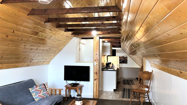 A cozy attic room with a wooden ceiling, a gray sofa, a small table, and a TV.
