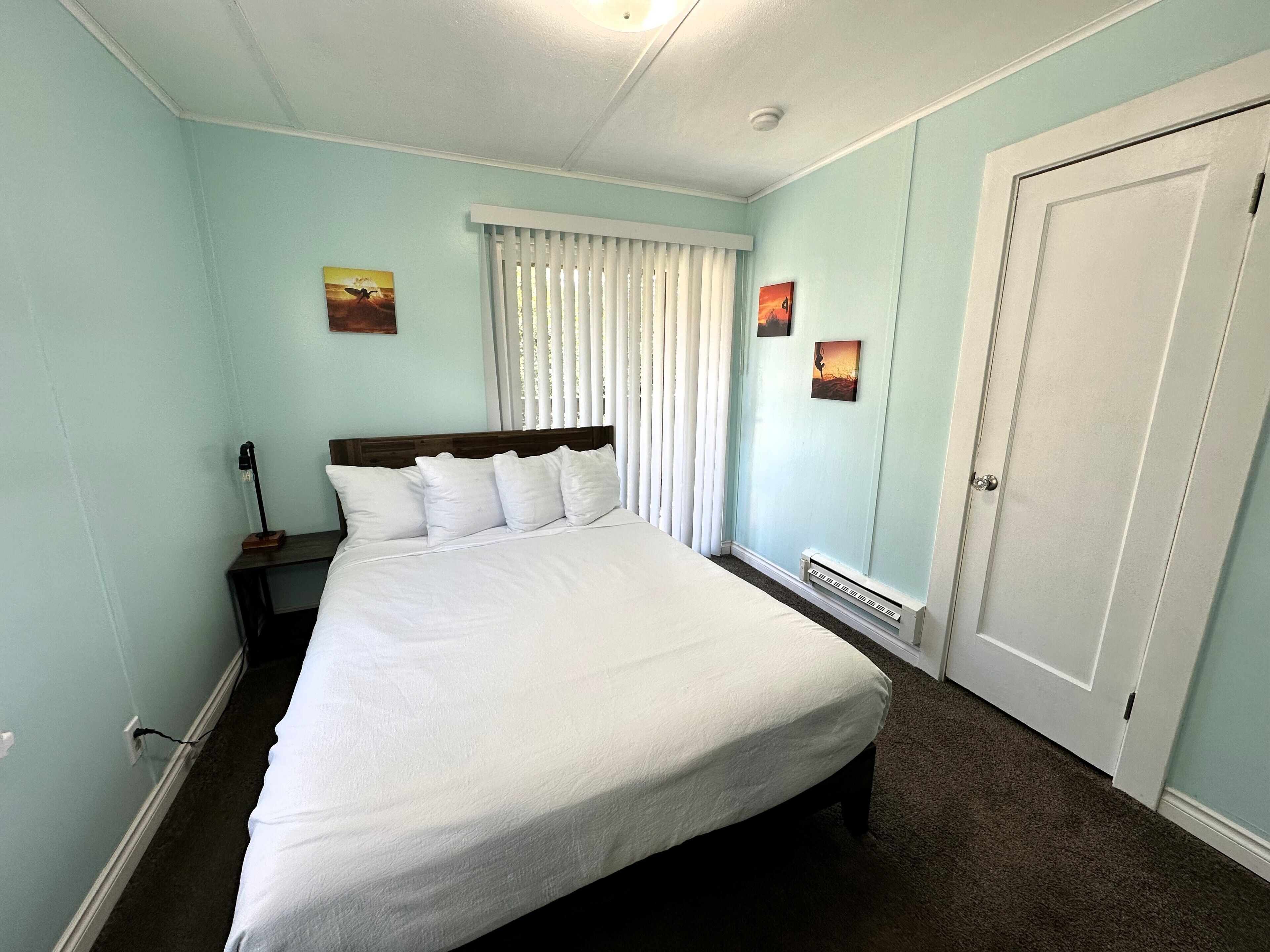 Una cama bien hecha en una habitación con paredes azul claro, una puerta blanca y dos cuadros pequeños en la pared.