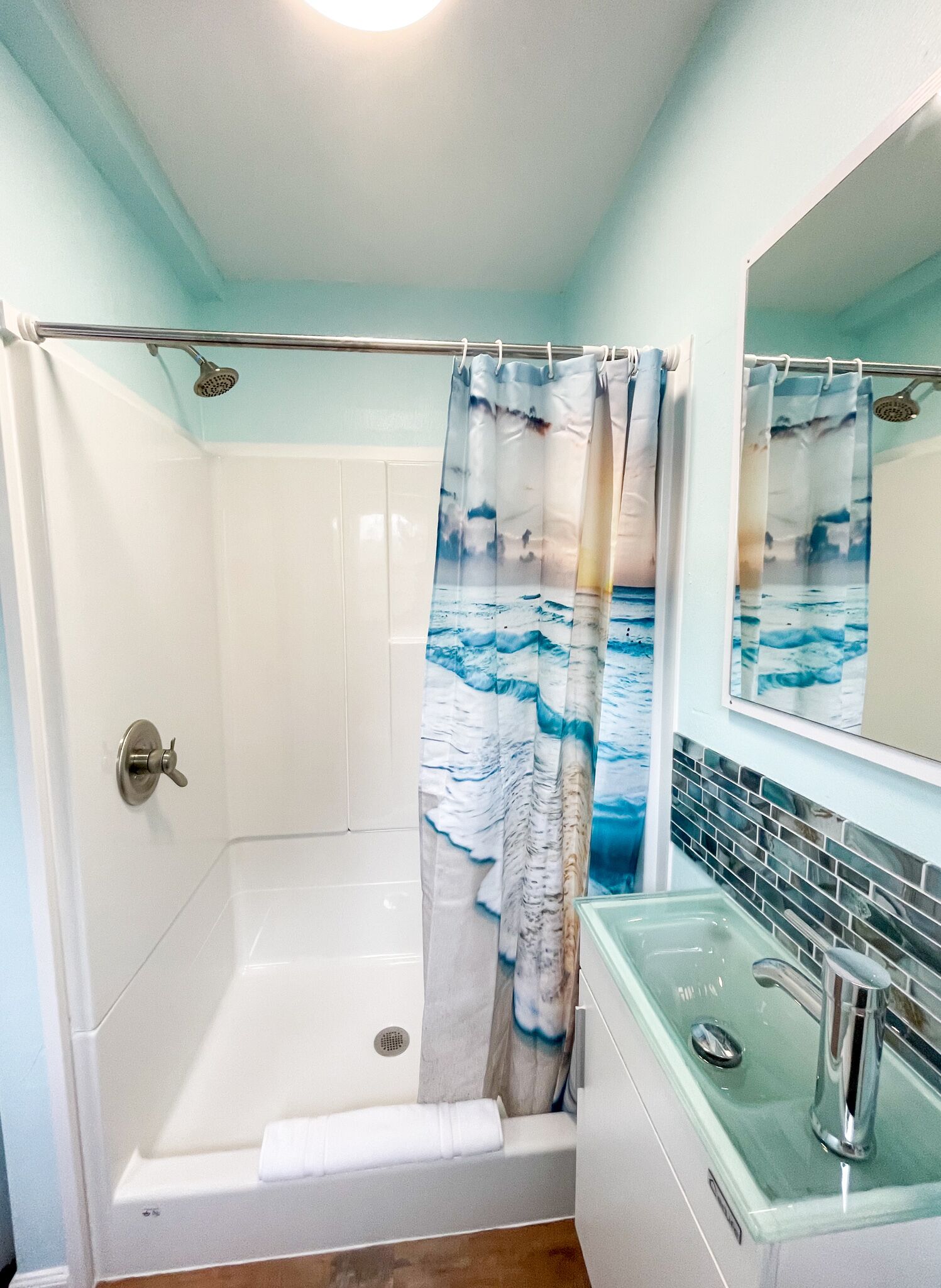 A bathroom with a shower curtain depicting ocean waves, a glass-enclosed bathtub, and a sink with a mirror above it.