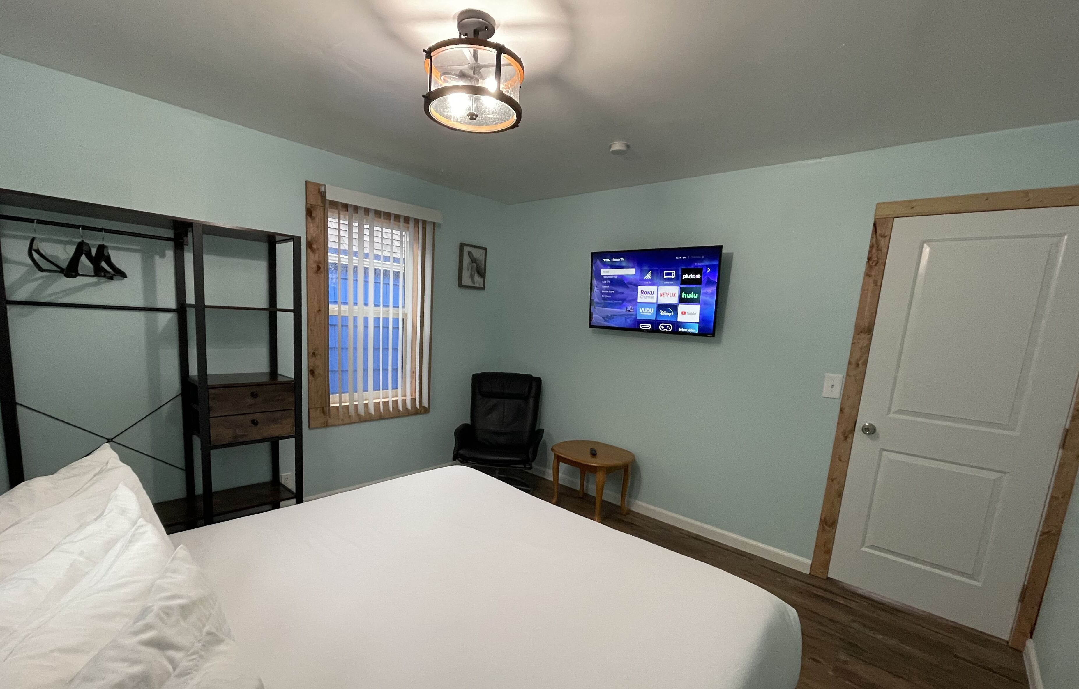A bedroom with a bed, a TV mounted on the wall, a wooden door, and a window with blinds.