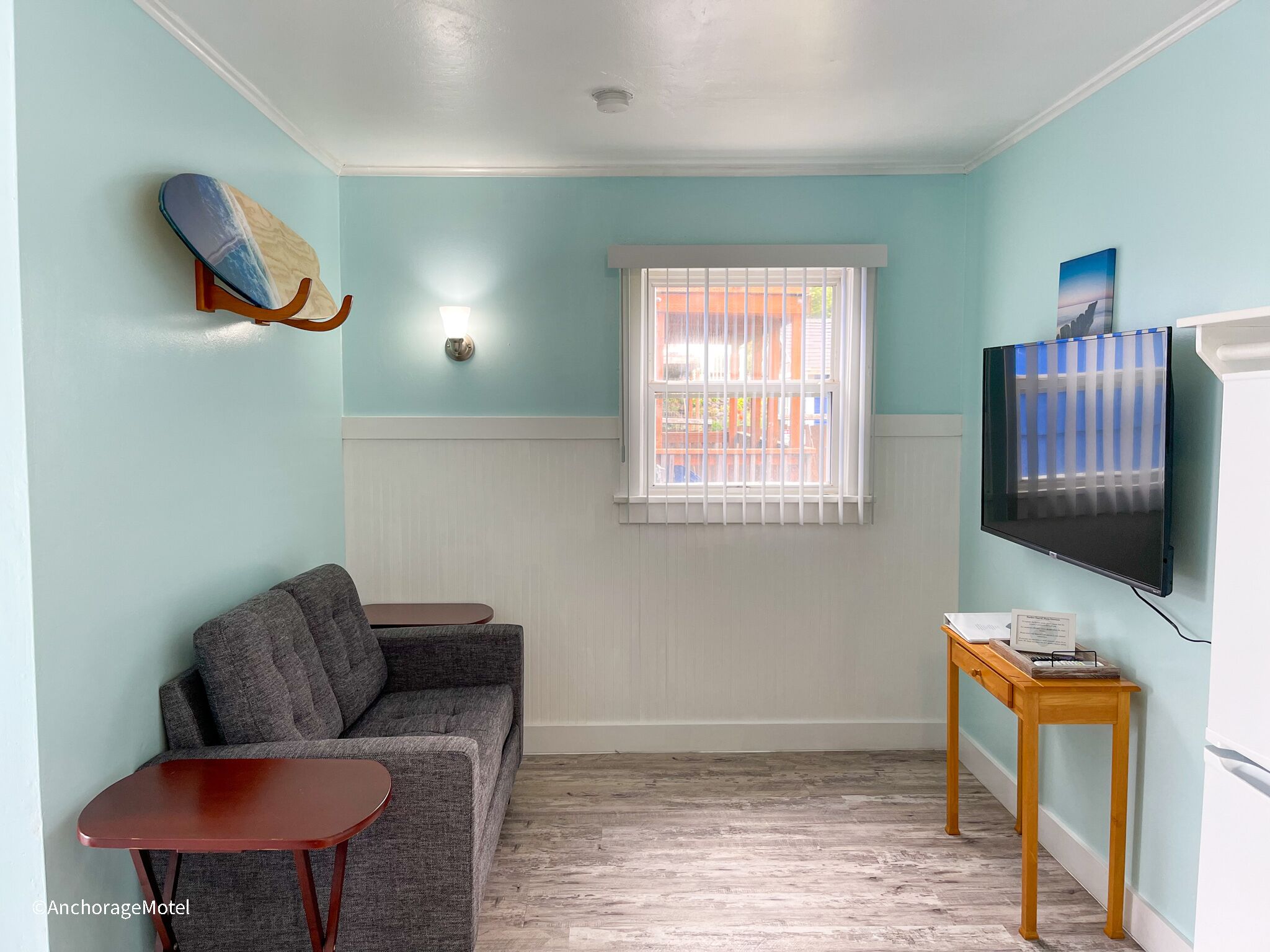 Een kamer met een grijze bank, een klein houten tafeltje, een aan de muur bevestigde televisie en een raam met witte zonwering.