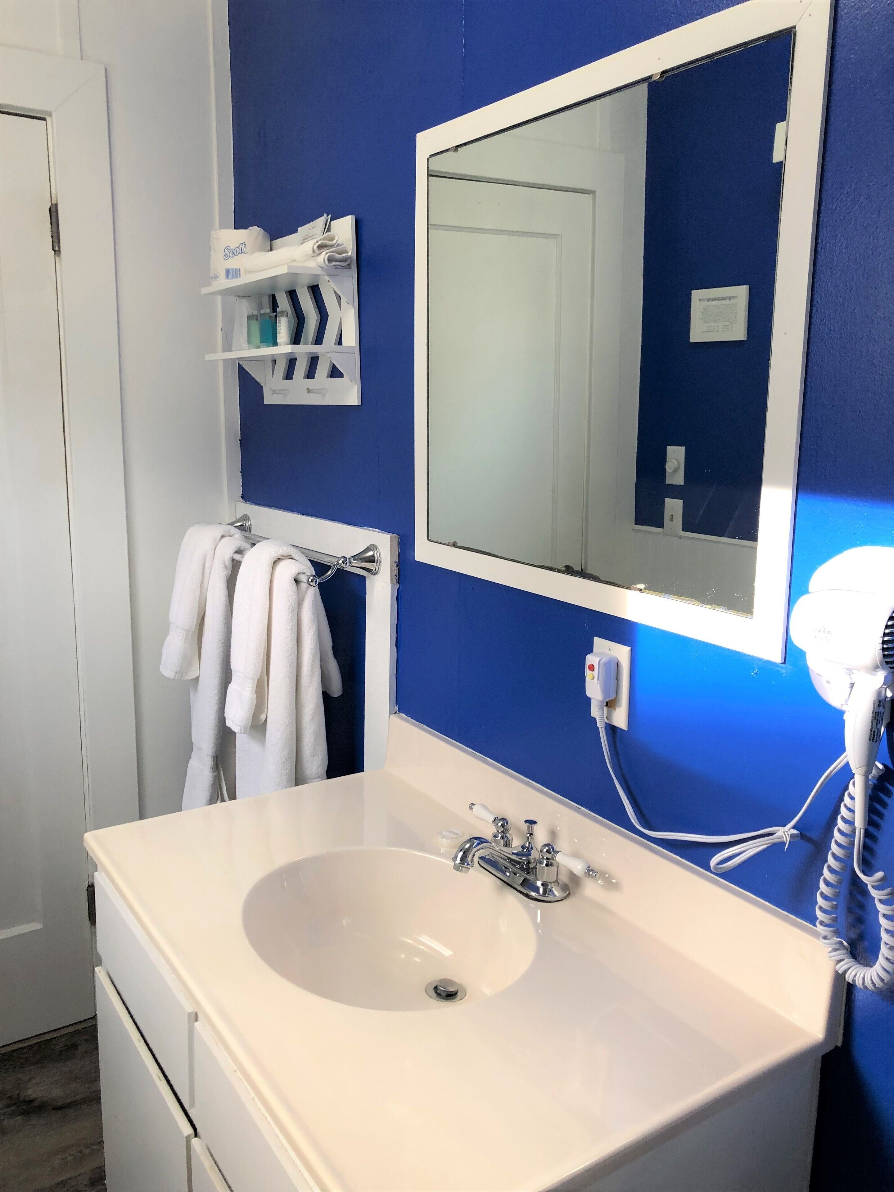 Une salle de bain avec un lavabo blanc, un miroir et un sèche-cheveux. Des serviettes sont suspendues sur un porte-serviettes et il y a une étagère avec des articles de toilette.