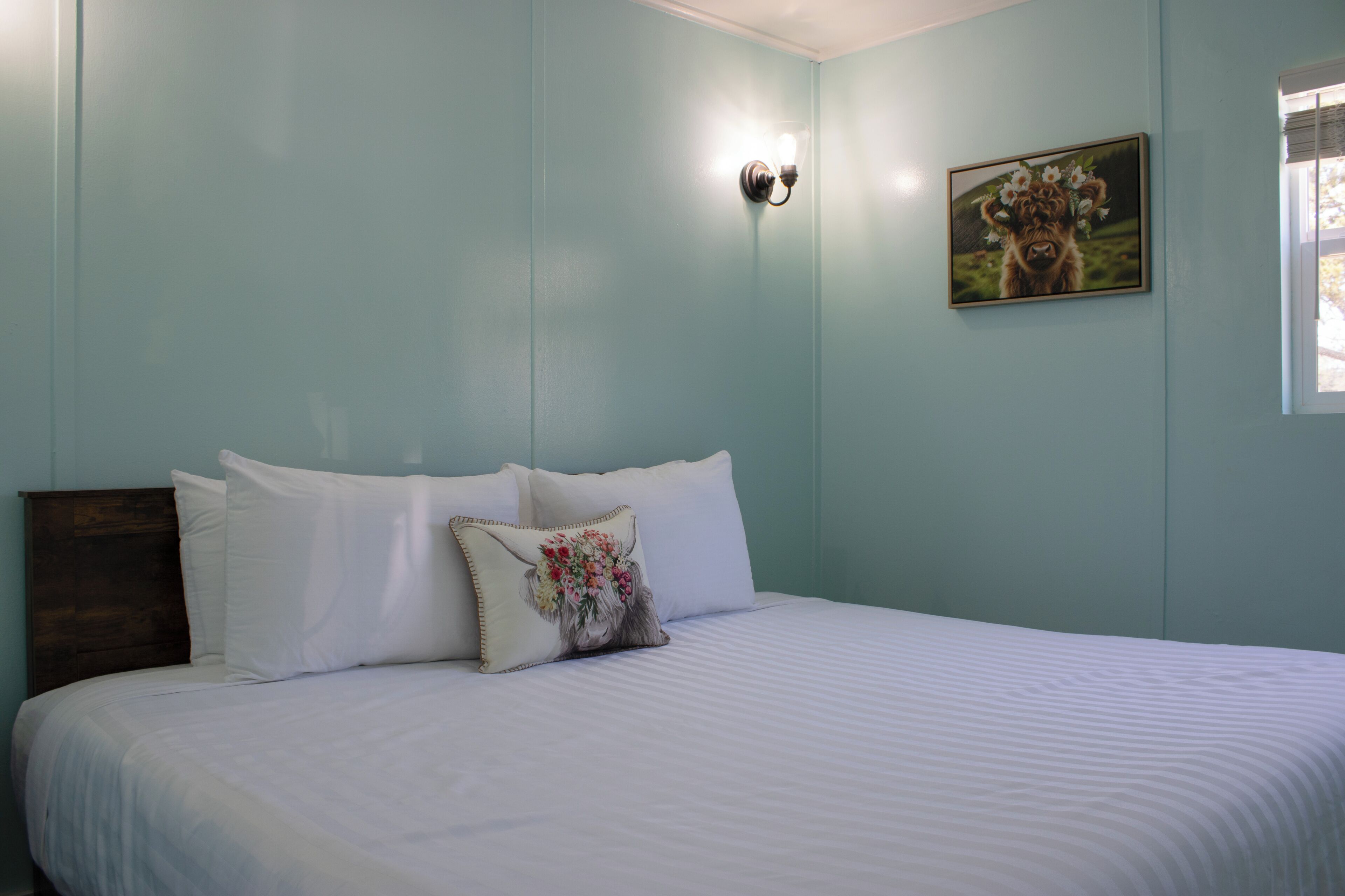 Un dormitorio con cama, ropa de cama blanca, un cojín decorativo, una lámpara de pared y un cuadro enmarcado.