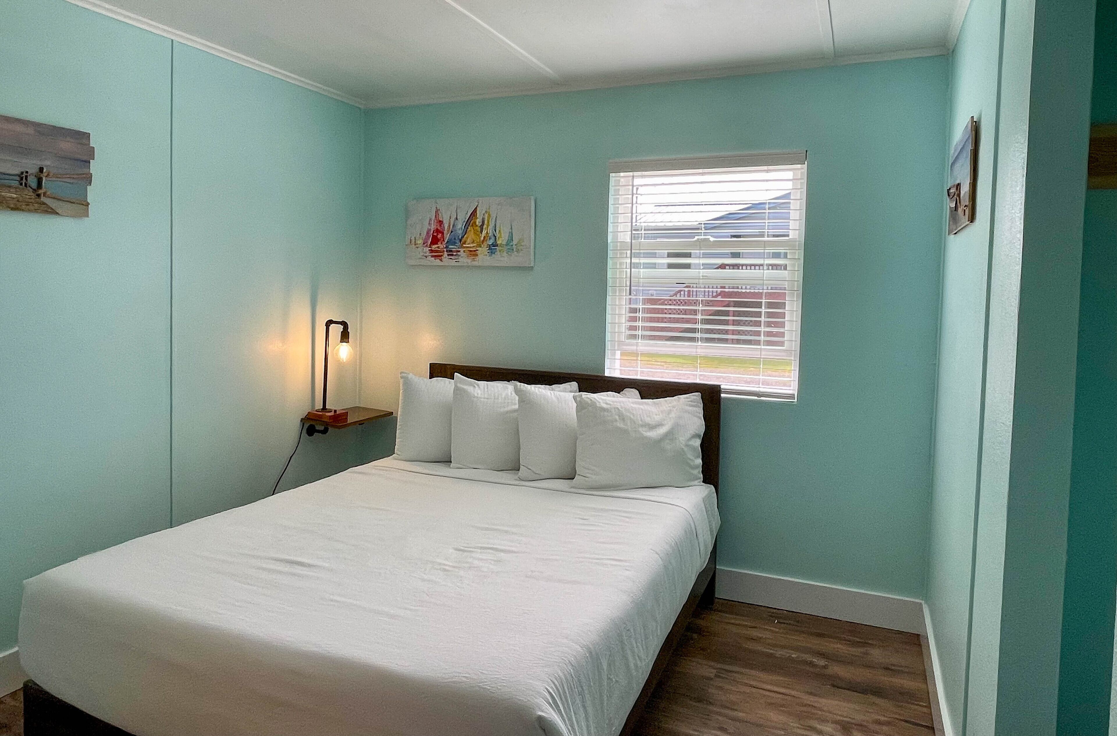 Un dormitorio con cama, una mesita de noche, una lámpara, un ventanal con persianas y un cuadro en la pared.