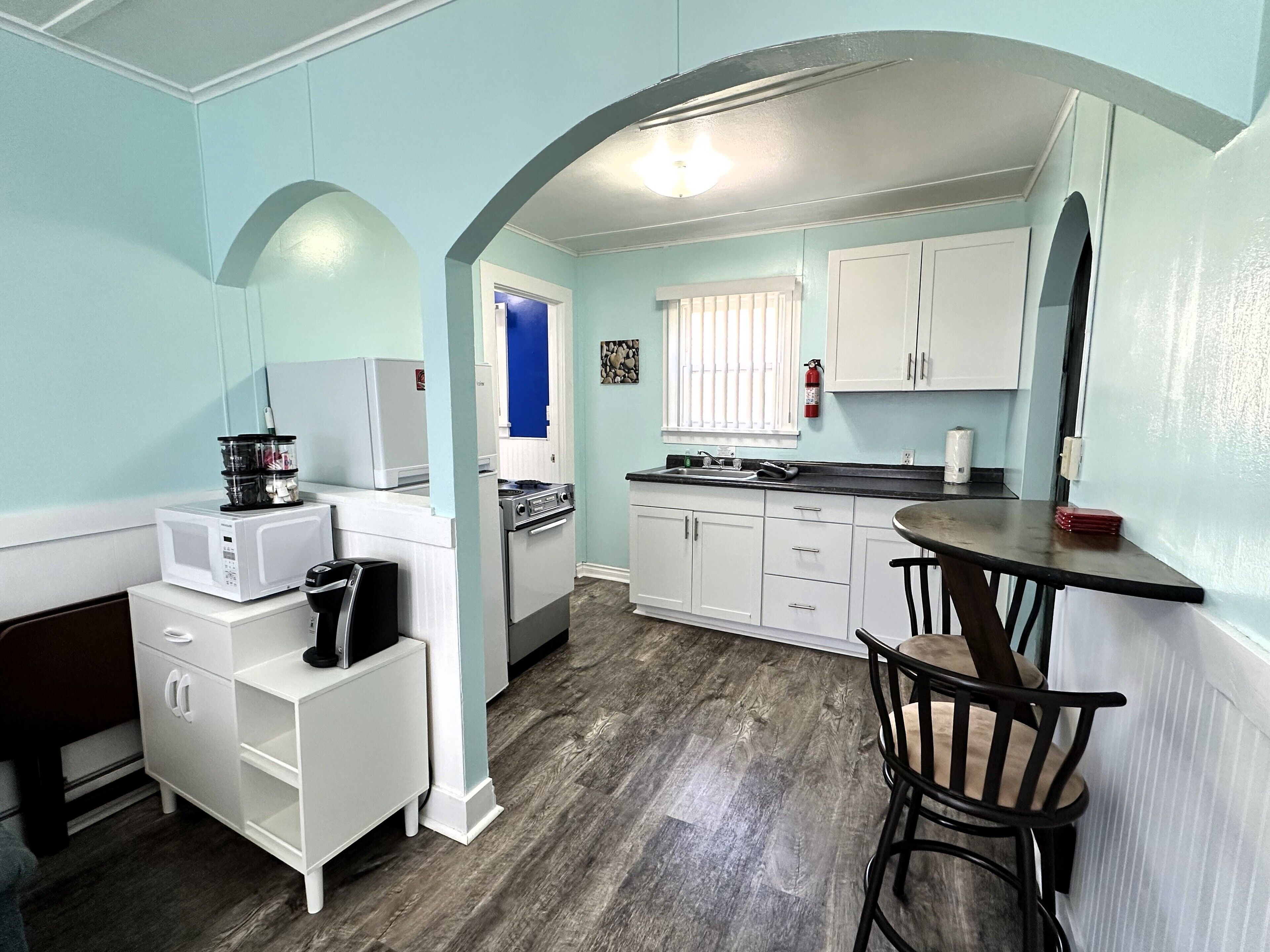 A compact kitchen with white cabinets, a microwave, a stove, and a sink. There is a small dining area with a table and chairs.