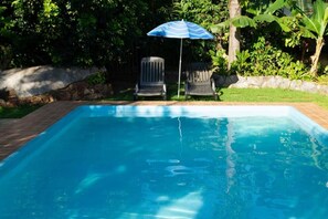 A rectangular swimming pool with a tiled edge, surrounded by a brick patio. There are two chairs and a striped umbrella near the pool. - Pousada Chão de Pedra (Bonito)