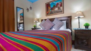 Habitación de hotel con una cama grande, cabecera de madera, manta a rayas coloridas, dos lámparas de noche, una pequeña planta en maceta y un cuadro en la pared.