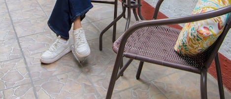Una persona sentada a una mesa redonda con una copa de bebida roja, leyendo un libro, en un espacio exterior con plantas en macetas y suelo de baldosas.