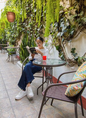 Una persona sentada a una mesa redonda con una copa de bebida roja, leyendo un libro, en un espacio exterior con plantas en macetas y suelo de baldosas.