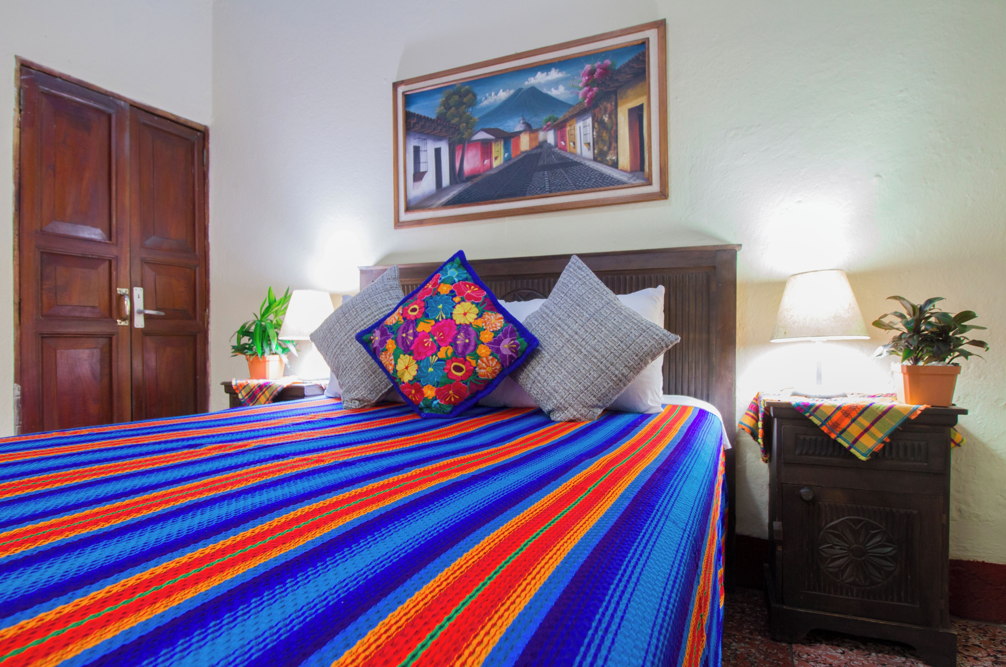 Uma cama com um edredom colorido listrado, duas almofadas, uma mesinha de cabeceira com abajur e um quadro na parede.