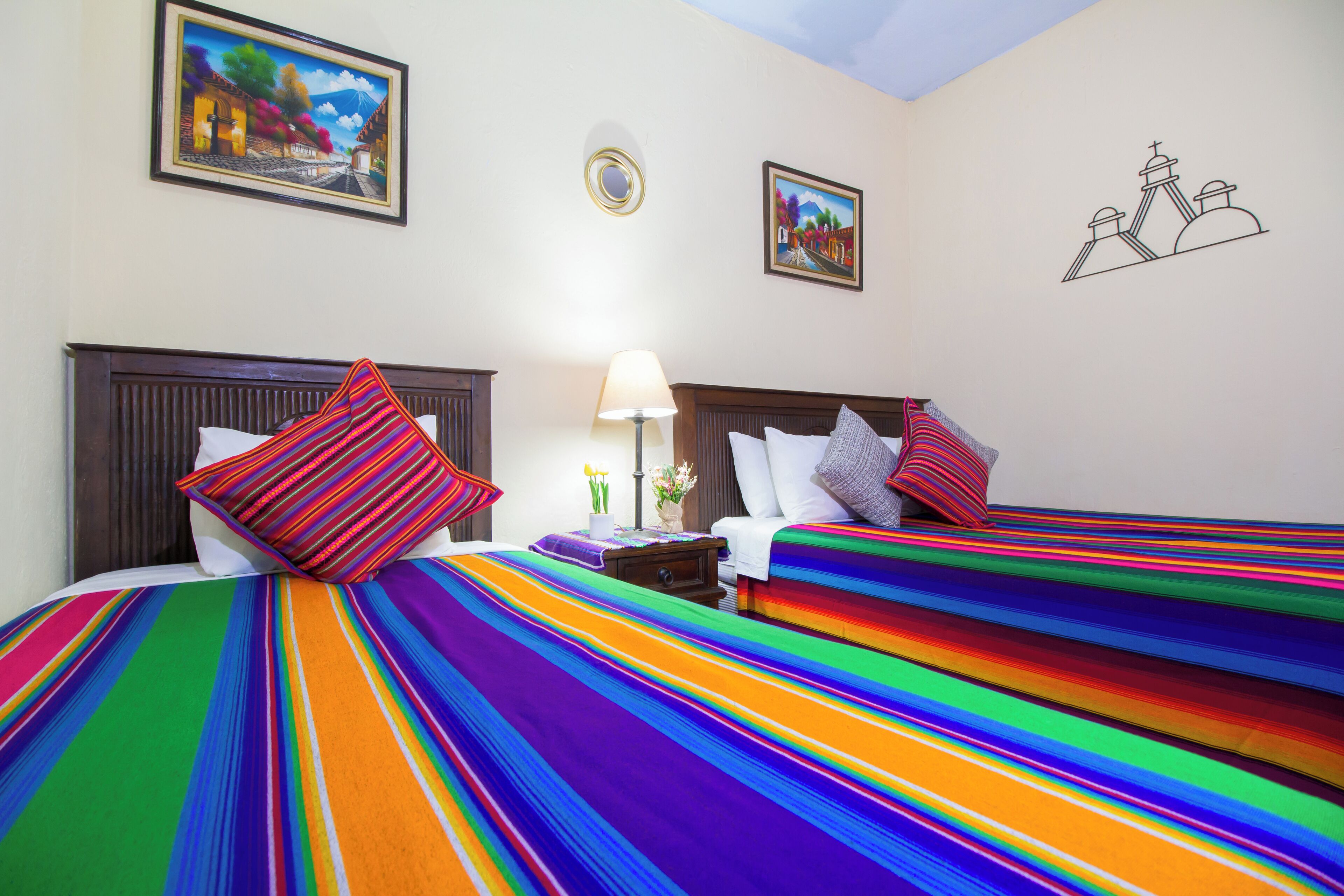 Quarto de hotel com duas camas, roupa de cama listrada e colorida, mesa de cabeceira com abajur e duas fotos emolduradas na parede.