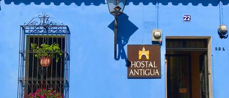 Un edificio azul con un letrero que dice 'Hostal Antigua' y un perro sentado afuera.