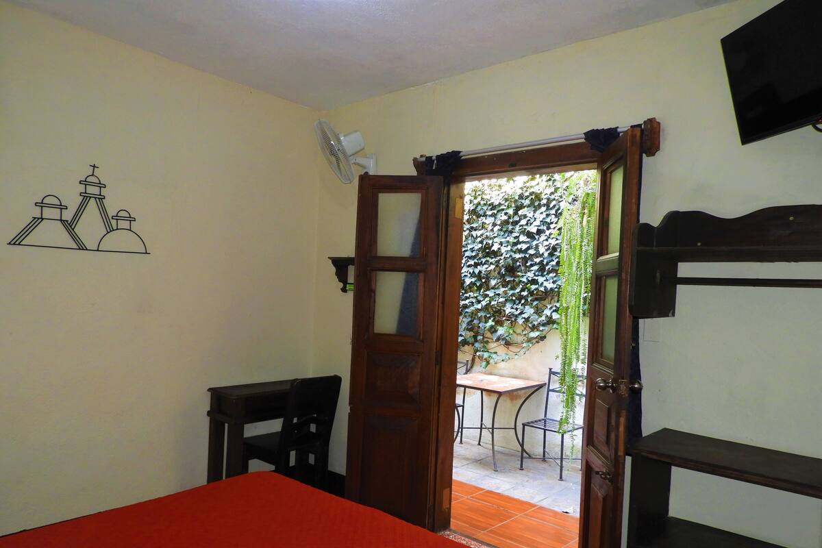 A room with a bed, a desk, a chair, a TV, and a door leading to an outdoor area with a table and chairs.