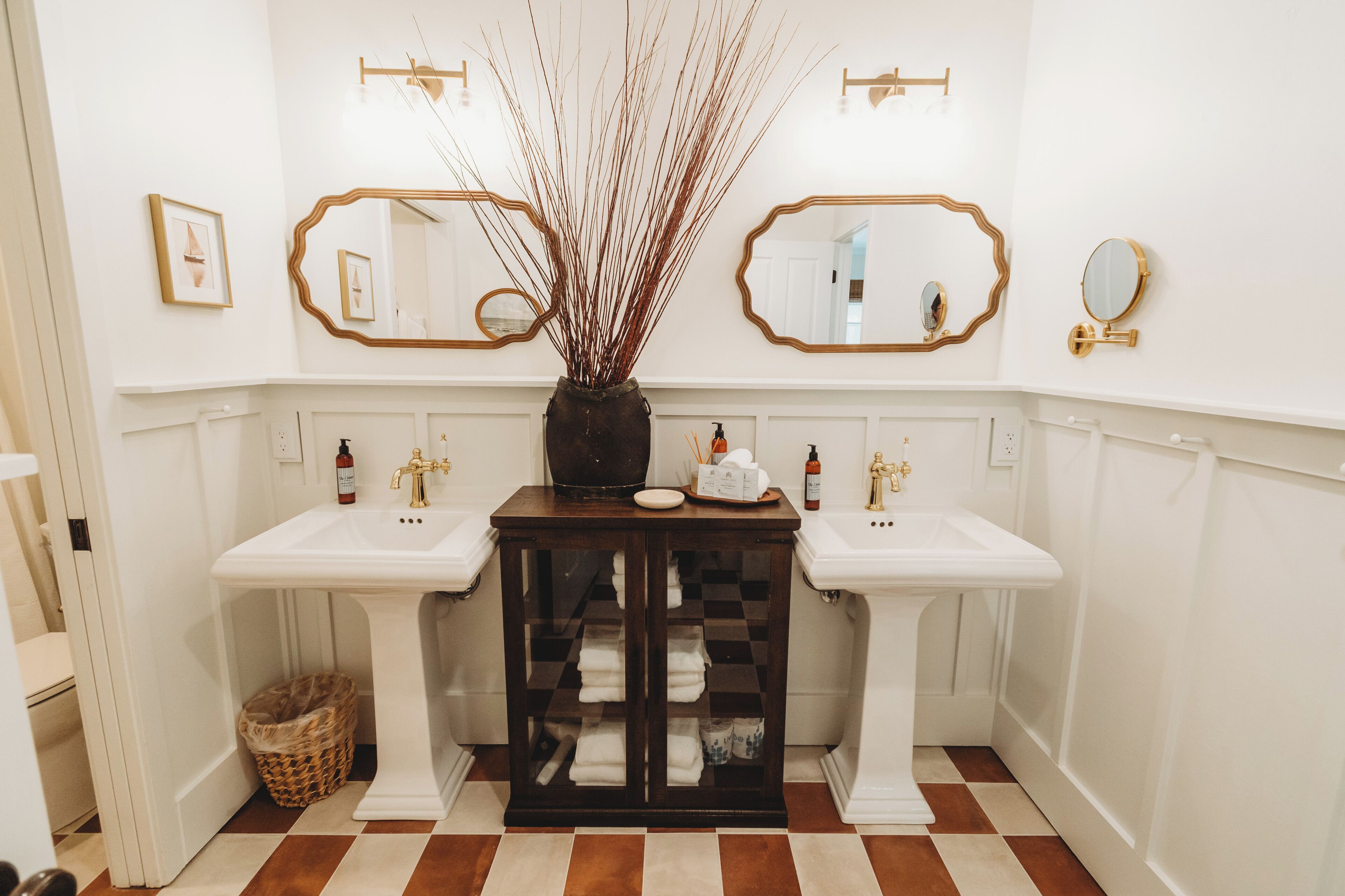 A bathroom with two sinks, a mirror, a vase with branches, and a cabinet with towels.