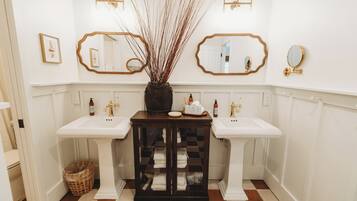 A bathroom with two sinks, a mirror, a vase with branches, and a cabinet with towels.