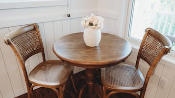 Ein runder Holztisch mit zwei Korbstühlen, einer weißen Vase mit Blumen und einem Fenster mit weißen Rollläden.