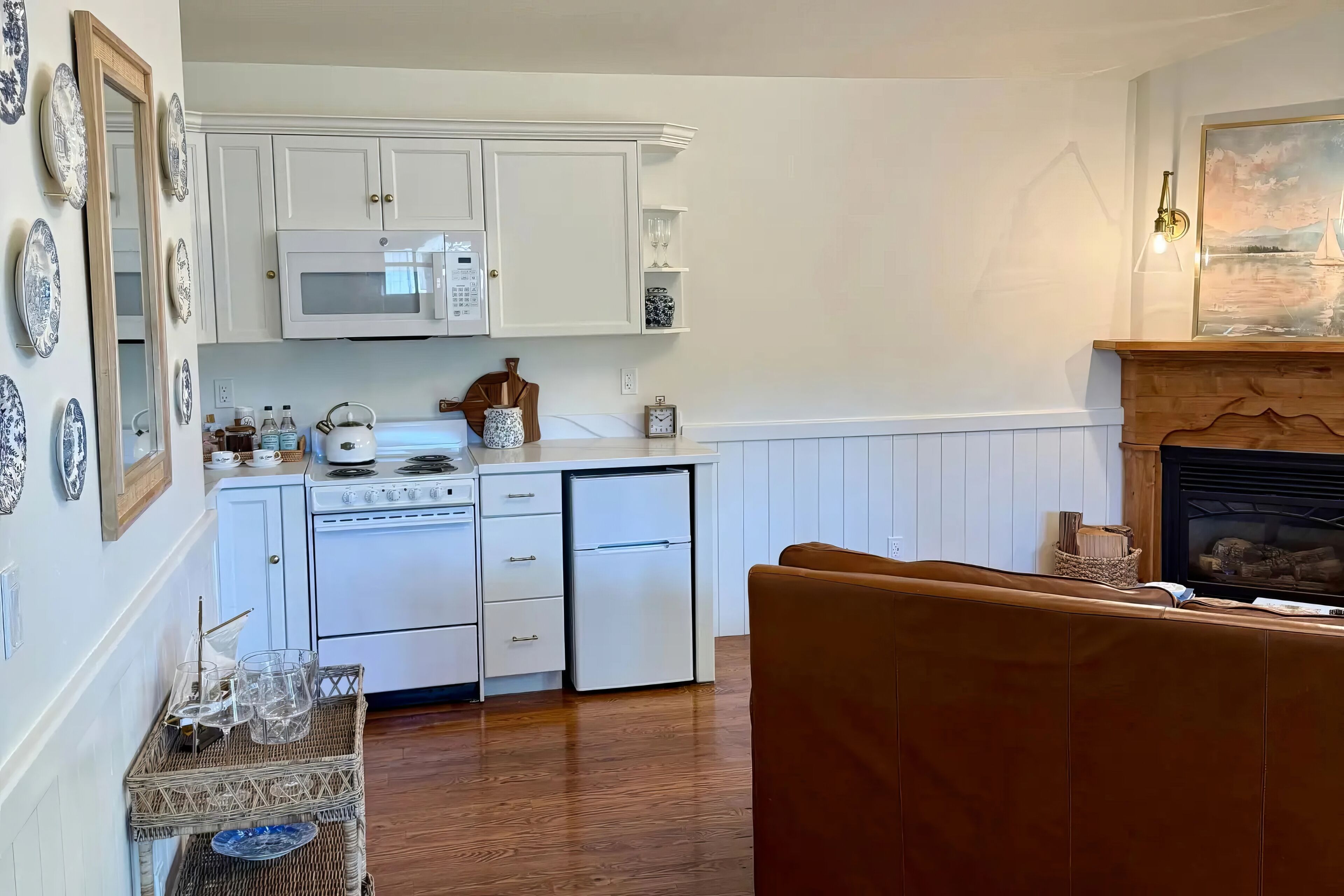Cocina con armarios blancos, microondas, estufa y refrigerador. Hay un banco de madera y una chimenea con un cuadro encima.