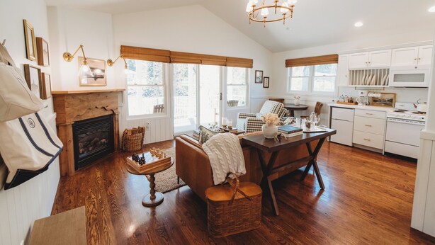 A spacious living area with a fireplace, wooden flooring, a dining table, and a kitchen with white cabinets. - The Cottages at Bigfork (Bigfork)