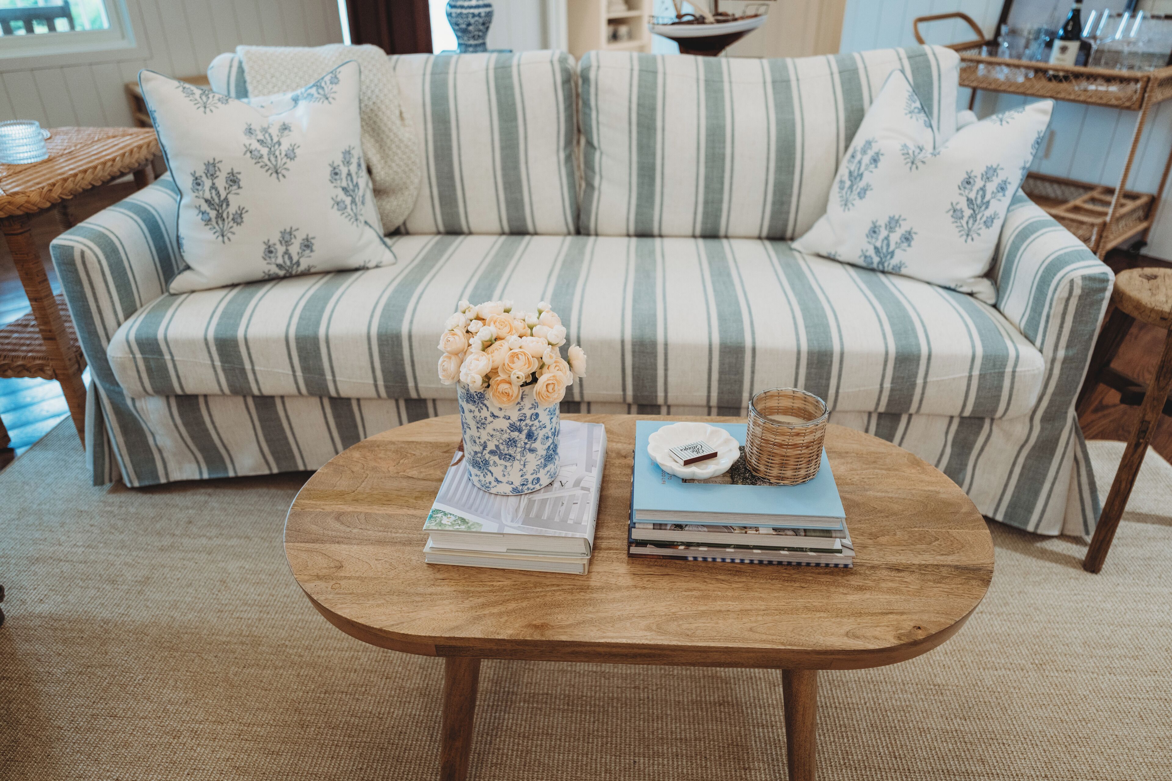 A striped sofa with floral pillows, a round wooden coffee table with books and a vase of flowers, and a wicker basket.