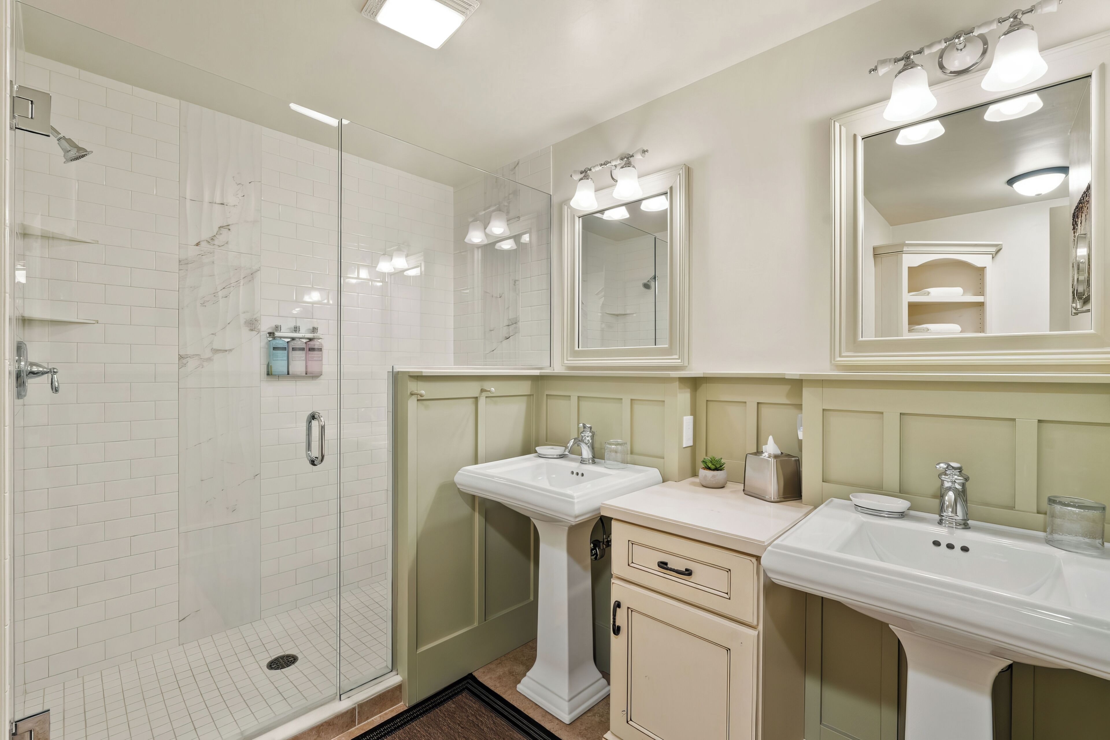 A bathroom with a glass-enclosed shower, two sinks, a mirror, and a vanity with drawers.