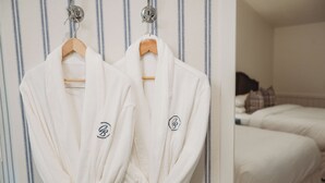 Two white robes hanging on wooden hangers against a striped wall, with a bed and pillows in the background. - The Cottages at Bigfork (Bigfork)