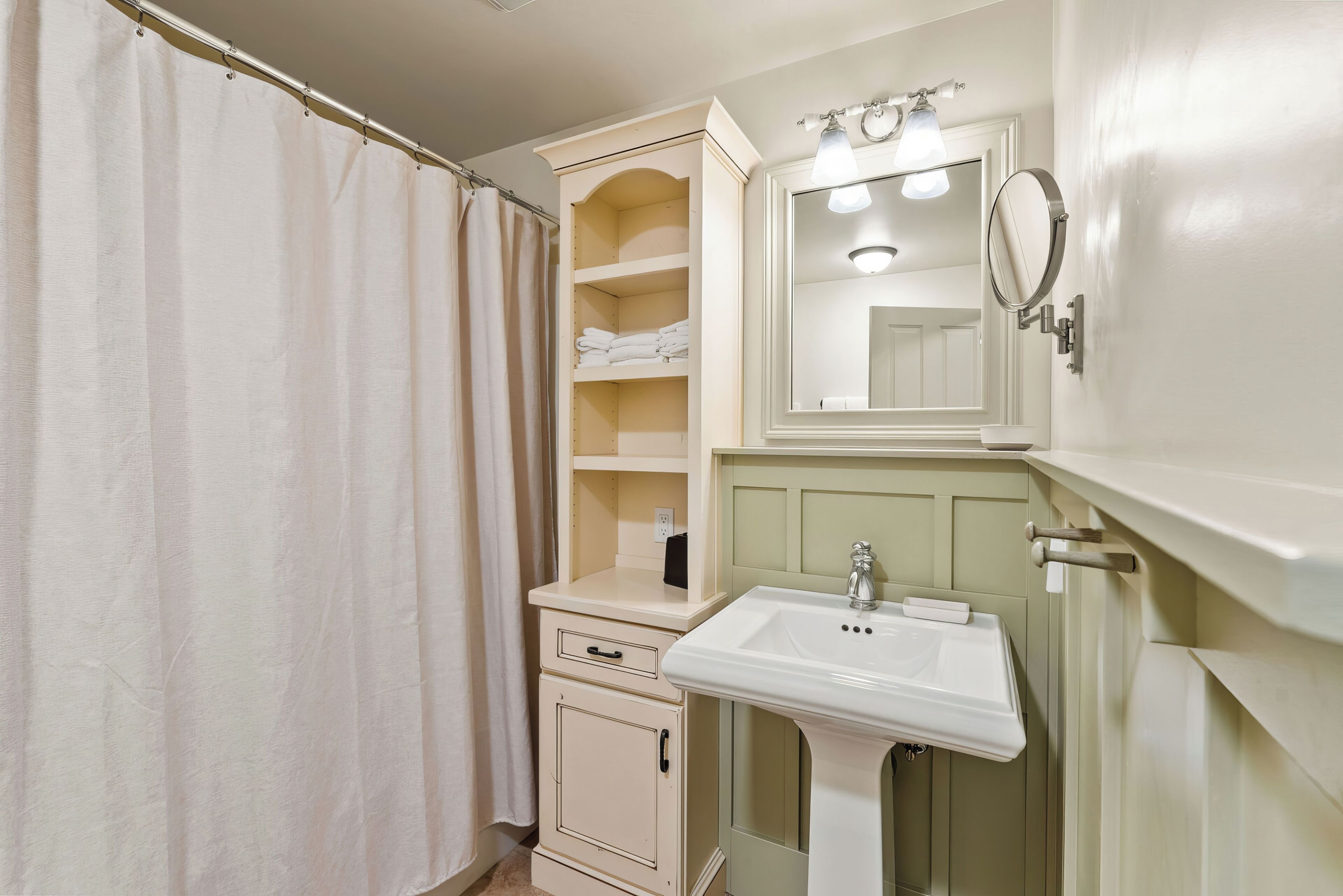 A bathroom with a shower curtain, a sink, a mirror, and a shelf with folded towels.