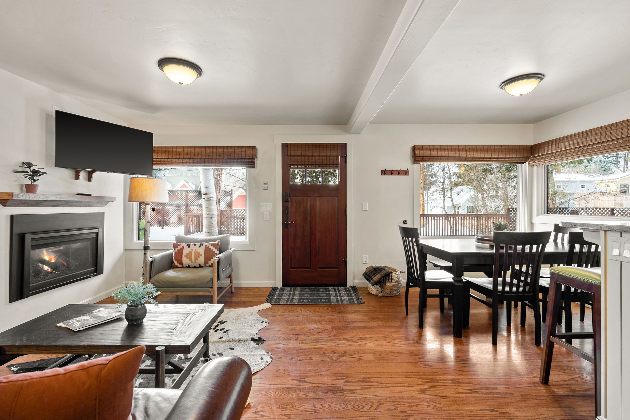 A living room with a fireplace, a television, a dining area with a table and chairs, and large windows with blinds.