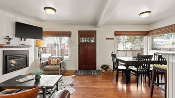 A living room with a fireplace, a television, a dining area with a table and chairs, and large windows with blinds.