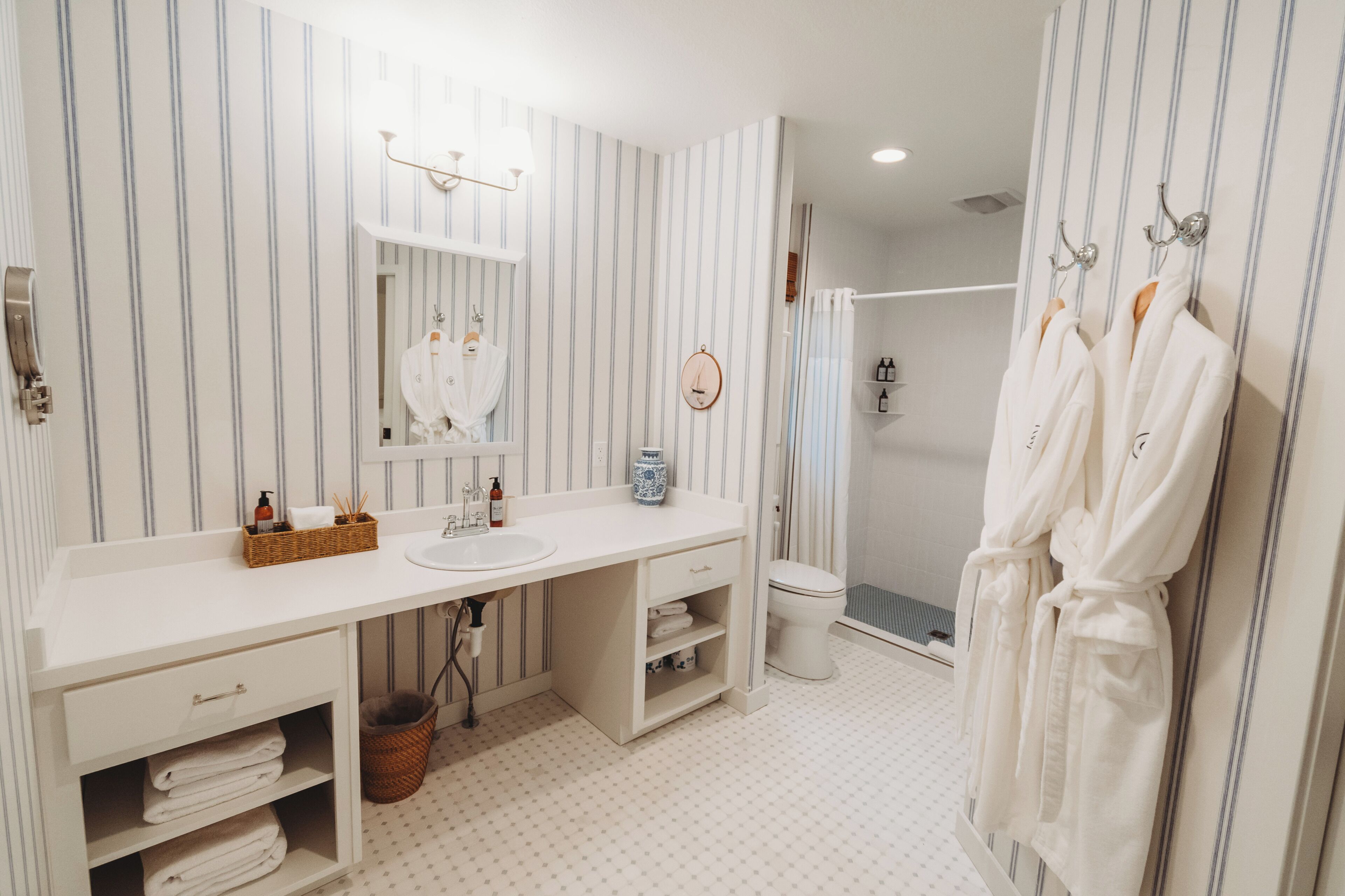 Une salle de bain avec un papier peint à rayures, un lavabo blanc, un miroir, une douche et deux peignoirs blancs suspendus à des crochets.