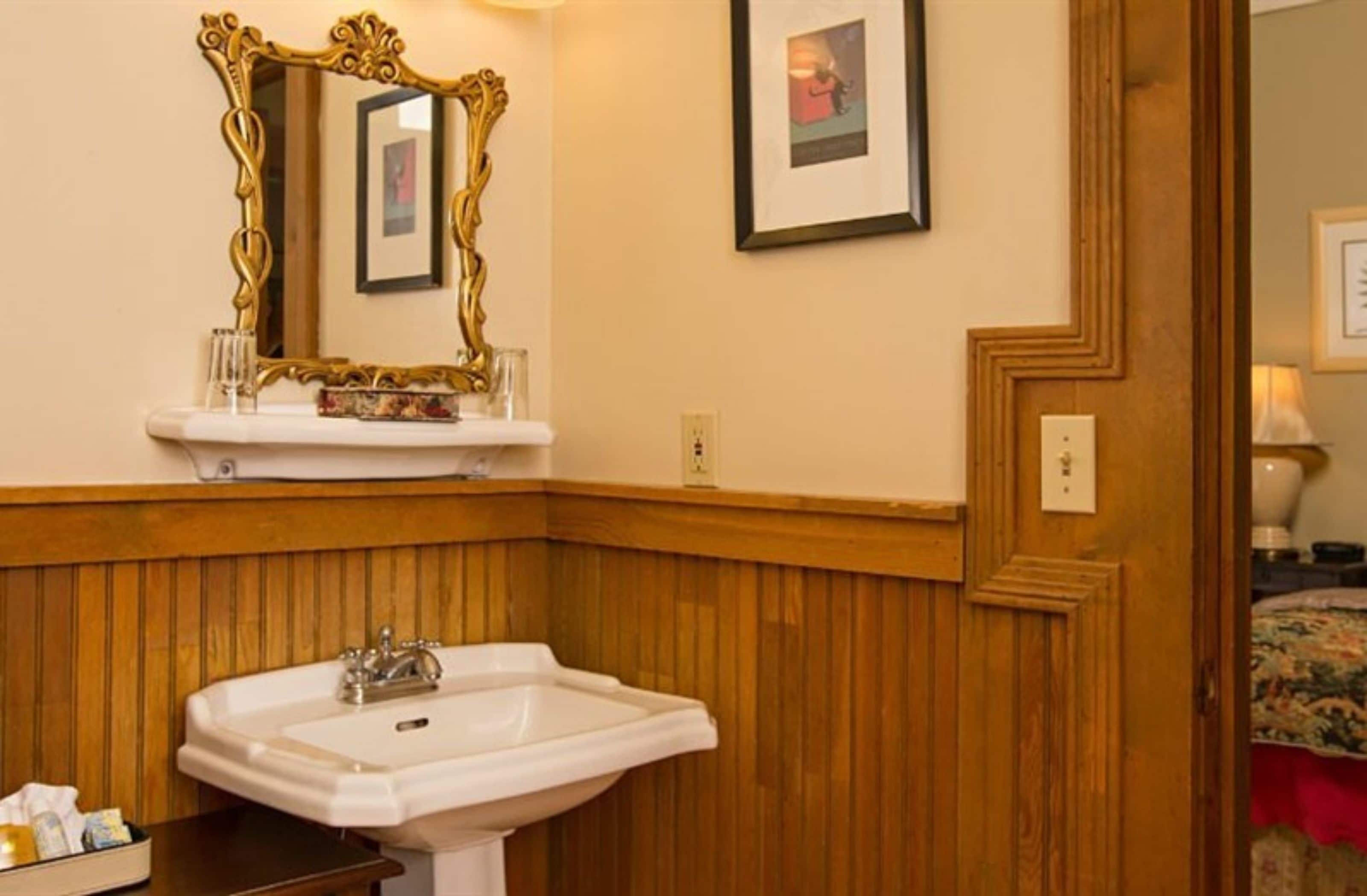A corner bathroom with a white sink, wooden paneling, a gold-framed mirror, and framed pictures on the wall.