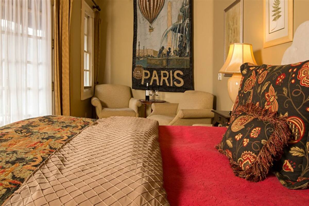 A hotel room with a bed, two chairs, a lamp, a wall with a Paris-themed poster, and a window with curtains.