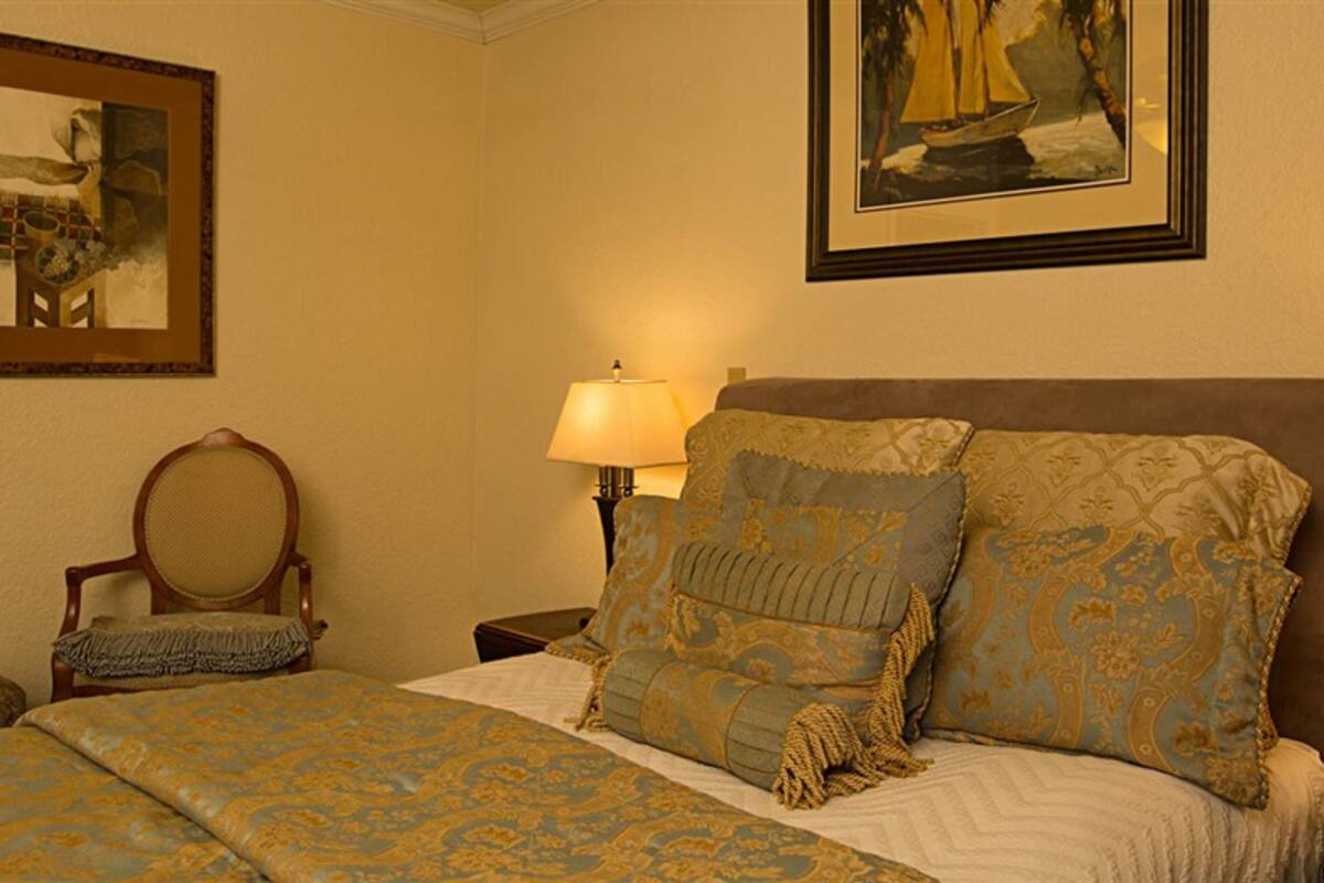 A hotel room with a bed, a chair, a nightstand with a lamp, and two framed artworks on the walls.