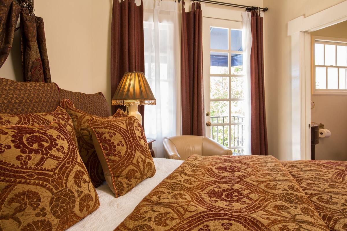A bedroom with a large bed, patterned pillows, a bedside lamp, a chair, and a window with curtains.